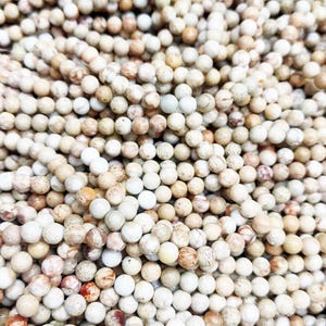 以下が含まれることがあります： クリーム、ベージュ、茶色の丸いビーズの山をクローズアップ。ビーズは斑点模様で、赤茶色とオレンジ色のマーキングがあります。これらのビーズはおそらくジュエリー作りに使用されます。