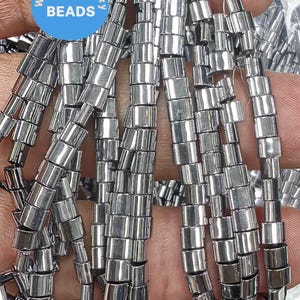 Può includere: Una collana di perline metalliche color argento, ogni perlina è un cilindro con un bordo leggermente arrotondato. Le perline sono disposte in un mucchio sciolto, con alcune perline che si sovrappongono.