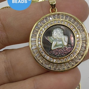 Puede incluir: Pendiente de oro con un camafeo redondo de nácar de abulón iridiscente que representa a un ángel con alas. El camafeo está rodeado por un círculo de piedras de circonita cúbica transparentes.