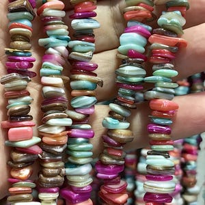 Könnte beinhalten: Nahaufnahme von Strängen aus farbigen, flachen Muschelkernperlen in Rosa-, Orange-, Blau- und Brauntönen. Die Perlen sind unregelmäßig geformt und aufgefädelt. Das Bild enthält einen blauen Kreis mit dem Text "WK DIY JEWERLY BEADS".