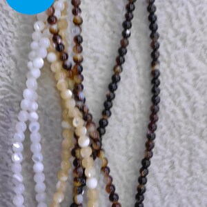 以下が含まれることがあります： 白、クリーム、茶色の丸いビーズが3本、わずかに虹色の光沢があります。ビーズはコードに糸に通され、緩やかな流れのあるパターンで配置されています。