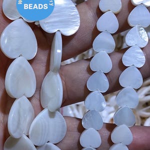May include: A strand of white heart-shaped shell beads. The beads are strung on a white cord and are arranged in a pattern of alternating sizes. The beads are smooth and have a natural iridescent sheen.