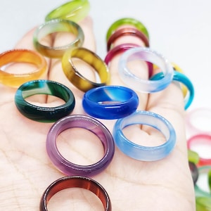 Op de afbeelding: Een verzameling kleurrijke edelsteenringen in verschillende tinten, waaronder groen, oranje, blauw, paars en rood. De ringen zijn glad en gepolijst en tonen de natuurlijke variaties in de steen. De ringen zijn in verschillende maten en stijlen.