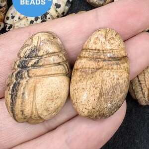 Op de afbeelding: Twee gegraveerde houten kralen in de vorm van een kever. De kralen zijn lichtbruin van kleur met donkerbruine strepen. De kralen zijn glad en gepolijst.