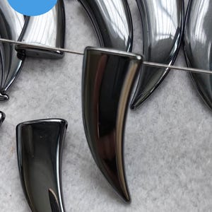 Peut inclure: Gros plan sur des perles noires brillantes en forme de corne. Les perles sont enfilées sur un fil argenté. Les perles mesurent environ 5 cm de long.