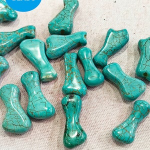 May include: A collection of turquoise-colored bone-shaped beads with brown veining. The beads have a smooth, polished surface and are arranged on a light-colored fabric. The image includes a blue circle with the word "BEADS".