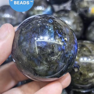 May include: A dark blue and green labradorite sphere with iridescent flashes of color. The sphere is smooth and polished, and it is being held in a hand.