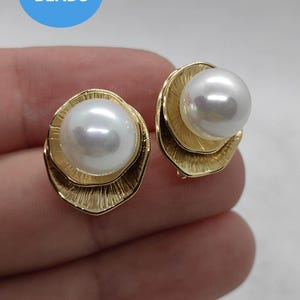 Op de afbeelding: Een paar goudkleurige oorstekers, elk met een grote, glanzende witte parel. De parels zijn gezet in een getextureerd, bladachtig gouden frame. Het woord "BEADS" staat in een blauwe cirkel linksboven.