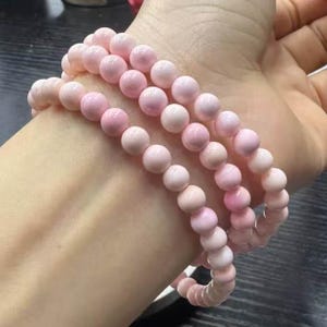 Puede incluir: Tres pulseras de cuentas rosadas con cuentas de tamaños graduados. Las cuentas son de color rosa claro y tienen un acabado liso y pulido.