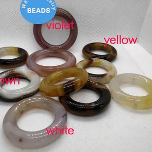 Puede incluir: Una colección de cuentas de piedra pulida en forma de rosquilla en varios colores, incluyendo violeta, marrón, amarillo y blanco. Las cuentas son circulares con un agujero en el centro. El texto "BEADS" está incluido.