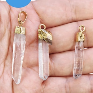 May include: Three clear quartz crystal pendants with gold-colored metal caps and loops. The crystals vary in size and shape, with one having a rough, natural edge. The pendants are displayed against a neutral background.