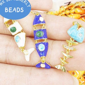 May include: Three fish-shaped jewelry charms with gold-tone metal frames. One charm is white enamel, one is blue enamel, and one is a light blue marbled enamel fish skeleton. Each features small inset stones and a loop for attachment at the top.