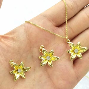 May include: A gold-colored jewelry set featuring a flower design. The set includes a necklace and two matching earrings. Each piece has a flower pendant with a small, clear stone in the center. The jewelry is displayed on a hand.