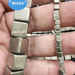 May include: Two strands of silver-toned pyrite cube beads. The beads are faceted and have a metallic sheen. The beads are strung on a wire and are approximately 1/2 inch in size.  The beads are displayed on a hand.