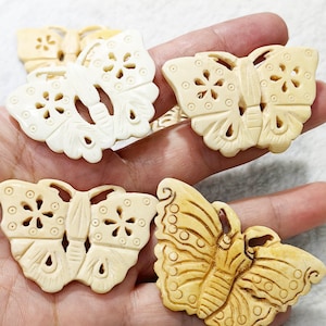 May include: Several carved butterfly ornaments in varying shades of cream and tan are displayed. The butterflies have intricate wing designs with cut-out patterns and detailed body features. The ornaments are held in a hand.