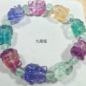 May include: A multi-colored bracelet with nine beads, each featuring a carved image of a mythical creature. The beads are made of fluorite, a crystal known for its iridescent properties. The bracelet is strung on a clear elastic cord.