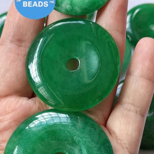 Puede incluir: Primer plano de una mano sosteniendo varias cuentas de jade verde en forma de rosquilla. Las cuentas tienen una superficie lisa y pulida y un agujero en el centro. Las cuentas son de color verde claro con vetas verde oscuro.