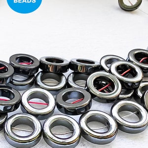 Puede incluir: Una hebra de cuentas de hematites negras con un agujero en el centro de cada cuenta. Las cuentas están dispuestas en un patrón circular.