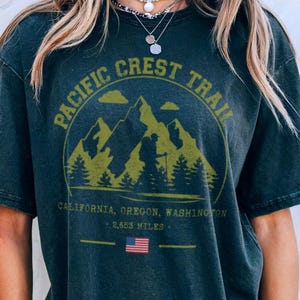 Op de afbeelding: Donkergrijs T-shirt met een cirkelvormige afbeelding met de tekst "Pacific Crest Trail" en een berglandschap in olijfgroen. Onder de afbeelding staan de woorden "California, Oregon, Washington" en "2.653 Miles". Een Amerikaanse vlag is ook zichtbaar.