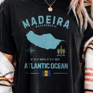 May include: Black t-shirt with a distressed print featuring the word "MADEIRA" above a map outline and the words "ATLANTIC OCEAN". Coordinates and a flag are also printed on the shirt. The shirt is black.