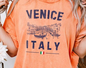 May include: Orange t-shirt with a vintage graphic of Venice, Italy. The design features the text "VENICE ITALY" above and below an illustration of a bridge and gondola. The Italian flag is below the text. The shirt has short sleeves.