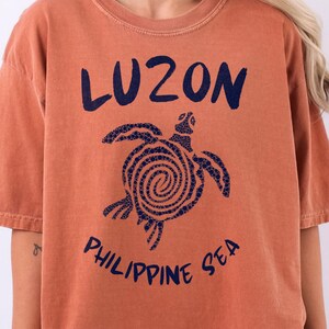 以下が含まれることがあります： 錆色の半袖Tシャツ。甲羅が渦巻き模様になったウミガメの濃い青色のグラフィックが描かれています。カメの上には「LUZON」、下には「PHILIPPINE SEA」という文字が濃い青色のインクでプリントされています。