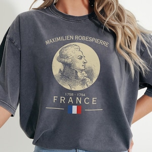 May include: Pepper-colored t-shirt featuring a portrait of Maximilien Robespierre, with the text "MAXIMILIEN ROBESPIERRE", "1758-1794", and "FRANCE" above a French flag graphic. The shirt has short sleeves and a relaxed fit.