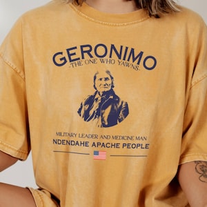 May include: Mustard-coloured t-shirt with the text "GERONIMO - THE ONE WHO YAWNS" above a portrait of Geronimo. Below the portrait, the text reads "MILITARY LEADER AND MEDICINE MAN NDENDAHE APACHE PEOPLE" with a small American flag.