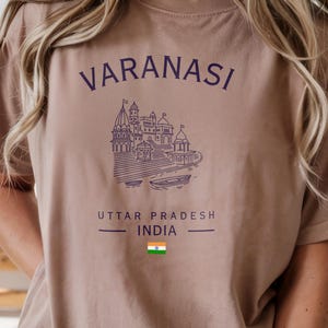 May include: A tan t-shirt with the word "VARANASI" in purple above a detailed purple illustration of a temple complex. Below the illustration, the text "UTTAR PRADESH INDIA" is printed, along with the Indian flag.
