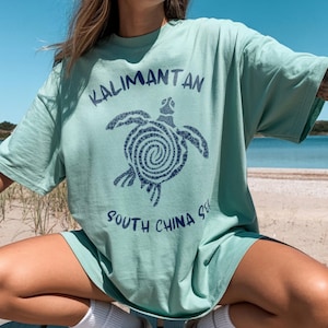 以下が含まれることがあります： アイランドリーフ色のTシャツで、渦巻き模様の甲羅を持つウミガメの濃い青色のグラフィックが描かれています。カメの上には「KALIMANTAN」、下には「SOUTH CHINA SEA」という文字が濃い青色のサンセリフ体でプリントされています。