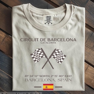 May include: Ivory-colored t-shirt with the text "CIRCUIT DE BARCELONA CATALUNYA" and crossed checkered flags. Below are coordinates and "BARCELONA, SPAIN" with the Spanish flag. The shirt is a Comfort Colors brand.
