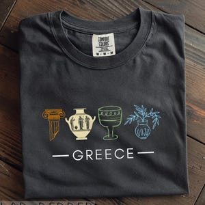 May include: Pepper-colored Comfort Colors t-shirt featuring a line drawing of Greek-inspired imagery. The design includes a column, vase, goblet, and vase with plants, with the word "GREECE" below. The shirt has a crew neck.