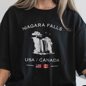 May include: Black t-shirt with the text "NIAGARA FALLS" above a graphic of the falls. Below the graphic, the text "USA / CANADA" is displayed with the American and Canadian flags. The shirt is black.