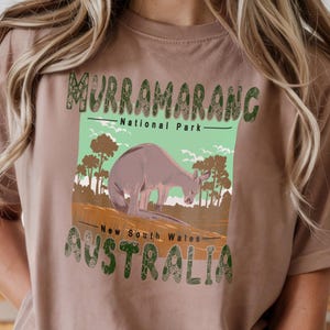 May include: Espresso-colored t-shirt featuring a graphic of a kangaroo in a natural landscape. The design includes the text "Murramarang National Park, New South Wales, Australia" in green and brown lettering. The shirt is a neutral color.
