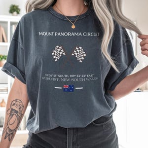 May include: Pepper-colored t-shirt featuring the text "MOUNT PANORAMA CIRCUIT" above two checkered flags. Below, coordinates and "BATHURST, NEW SOUTH WALES" are printed. An Australian flag is also visible. The shirt is a casual style.