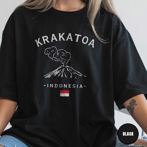 Peut inclure: T-shirt noir avec un dessin blanc d'un volcan en éruption, le mot "KRAKATOA" au-dessus et "-INDONESIA-" en dessous, avec le drapeau indonésien. Le t-shirt est de style décontracté.