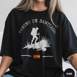 May include: Black t-shirt with a white graphic of a hiker with a backpack and walking stick, the text "CAMINO DE SANTIAGO" and the Spanish flag. The shirt is a casual style.