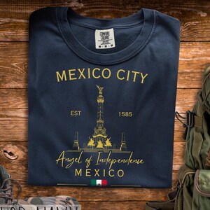 May include: Navy blue t-shirt featuring a gold graphic of the Angel of Independence monument in Mexico City. The text "MEXICO CITY" and "Angel of Independence" are printed above and below the monument. The shirt also features the text "EST 1585".