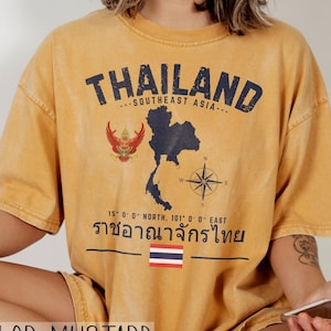 May include: Mustard-colored t-shirt with the word "THAILAND" in navy blue, along with a map of Thailand and the text "SOUTHEAST ASIA." Additional text and a compass rose are also present. The shirt also features the Thai flag.