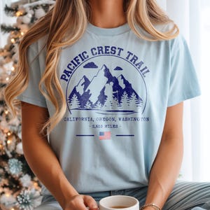 Op de afbeelding: Lichtblauw T-shirt met een marineblauwe afbeelding van bergen, bomen en de tekst "PACIFIC CREST TRAIL". Onder de afbeelding staan de woorden "CALIFORNIA, OREGON, WASHINGTON - 4265 km" en een Amerikaanse vlag.