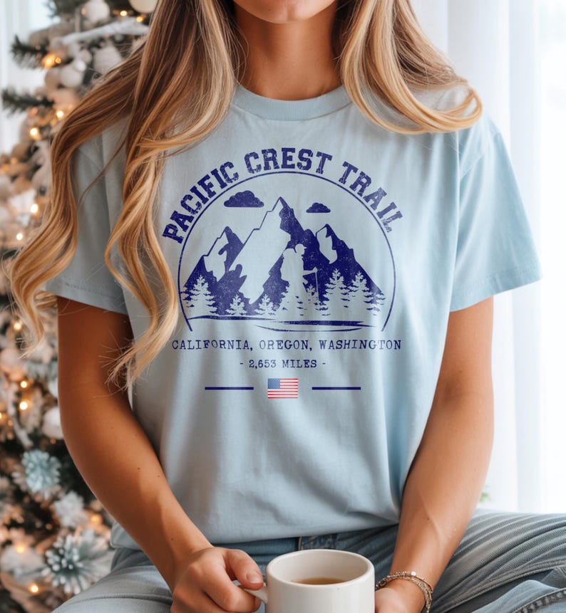 Op de afbeelding: Lichtblauw T-shirt met een marineblauwe afbeelding van bergen, bomen en de tekst "PACIFIC CREST TRAIL". Onder de afbeelding staan de woorden "CALIFORNIA, OREGON, WASHINGTON - 4265 km" en een Amerikaanse vlag.