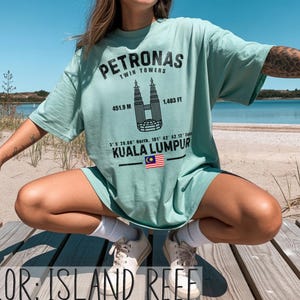 Peut inclure: Un t-shirt couleur r&eacute;cif insulaire avec un motif noir des tours Petronas de Kuala Lumpur. L'impression comprend le texte Petronas Twin Towers, 451,9 m, 1 483 ft, des coordonn&eacute;es, Kuala Lumpur et un petit drapeau malaisien en illustration.