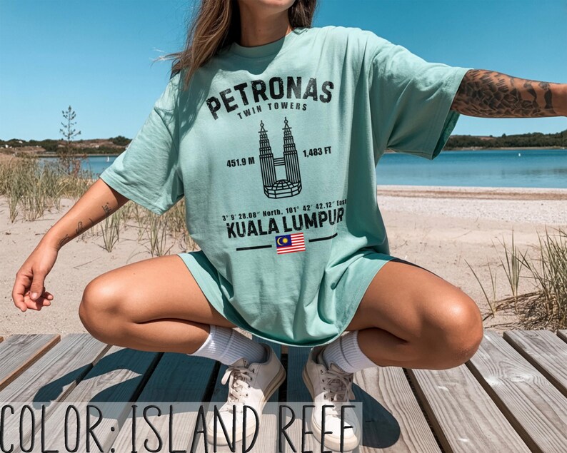 Peut inclure: Un t-shirt couleur r&eacute;cif insulaire avec un motif noir des tours Petronas de Kuala Lumpur. L'impression comprend le texte Petronas Twin Towers, 451,9 m, 1 483 ft, des coordonn&eacute;es, Kuala Lumpur et un petit drapeau malaisien en illustration.