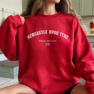 May include: A red sweatshirt with the text "NEWCASTLE UPON TYNE" in white arched lettering. Below the text are the words "GREAT BRITAIN" and a small Union Jack flag. The sweatshirt is a solid red color.
