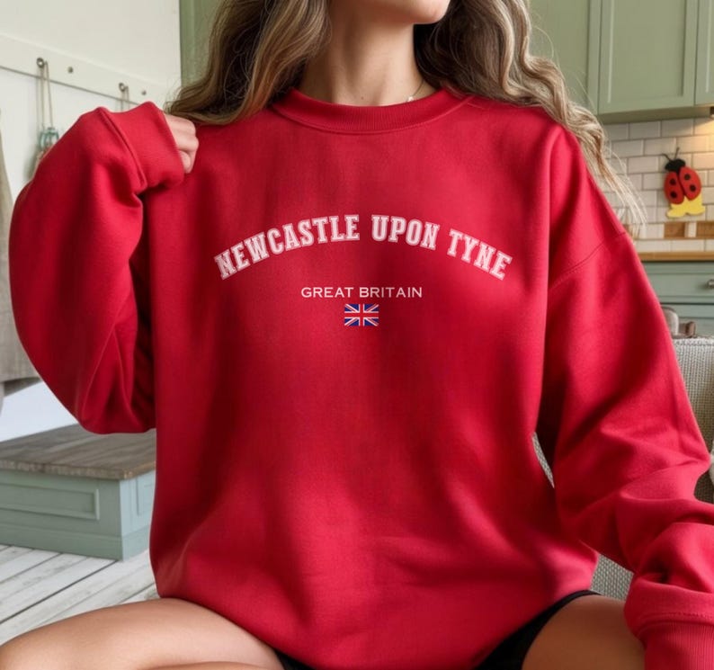 May include: A red sweatshirt with the text "NEWCASTLE UPON TYNE" in white arched lettering. Below the text are the words "GREAT BRITAIN" and a small Union Jack flag. The sweatshirt is a solid red color.