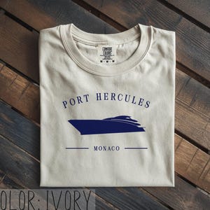 May include: Ivory-colored t-shirt with a navy blue boat graphic and the words "PORT HERCULES MONACO". The shirt is made of a soft material and has a crew neck. The shirt is folded on a wooden surface.