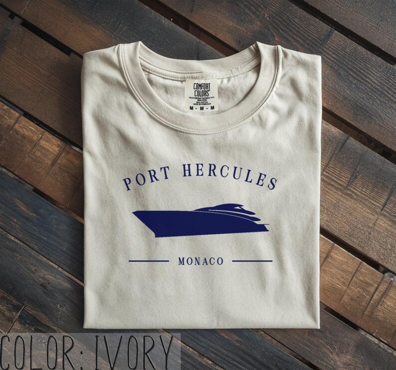 May include: Ivory-colored t-shirt with a navy blue boat graphic and the words "PORT HERCULES MONACO". The shirt is made of a soft material and has a crew neck. The shirt is folded on a wooden surface.