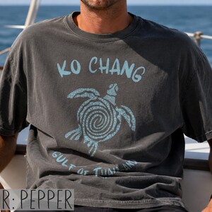 以下が含まれることがあります： ペッパーカラーの半袖Tシャツ。渦巻き模様の甲羅を持つウミガメのライトブルーのグラフィックが描かれています。カメの上には「KO CHANG」、下には「GULF OF THAILAND」という文字がライトブルーでプリントされています。