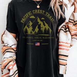 Op de afbeelding: Zwart T-shirt met een cirkelvormige afbeelding van bergen, bomen en de tekst "Pacific Crest Trail". Daaronder staat "California, Oregon, Washington - 2.653 mijl" met een kleine Amerikaanse vlag.