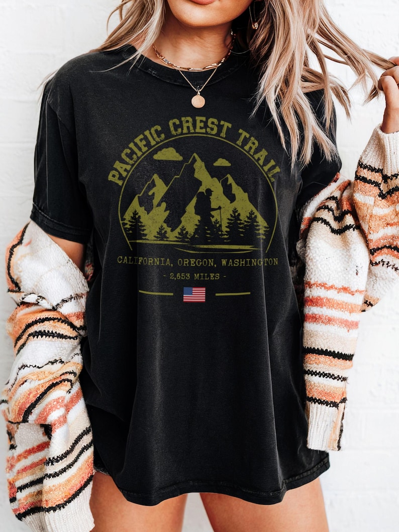 Op de afbeelding: Zwart T-shirt met een cirkelvormige afbeelding van bergen, bomen en de tekst "Pacific Crest Trail". Daaronder staat "California, Oregon, Washington - 2.653 mijl" met een kleine Amerikaanse vlag.